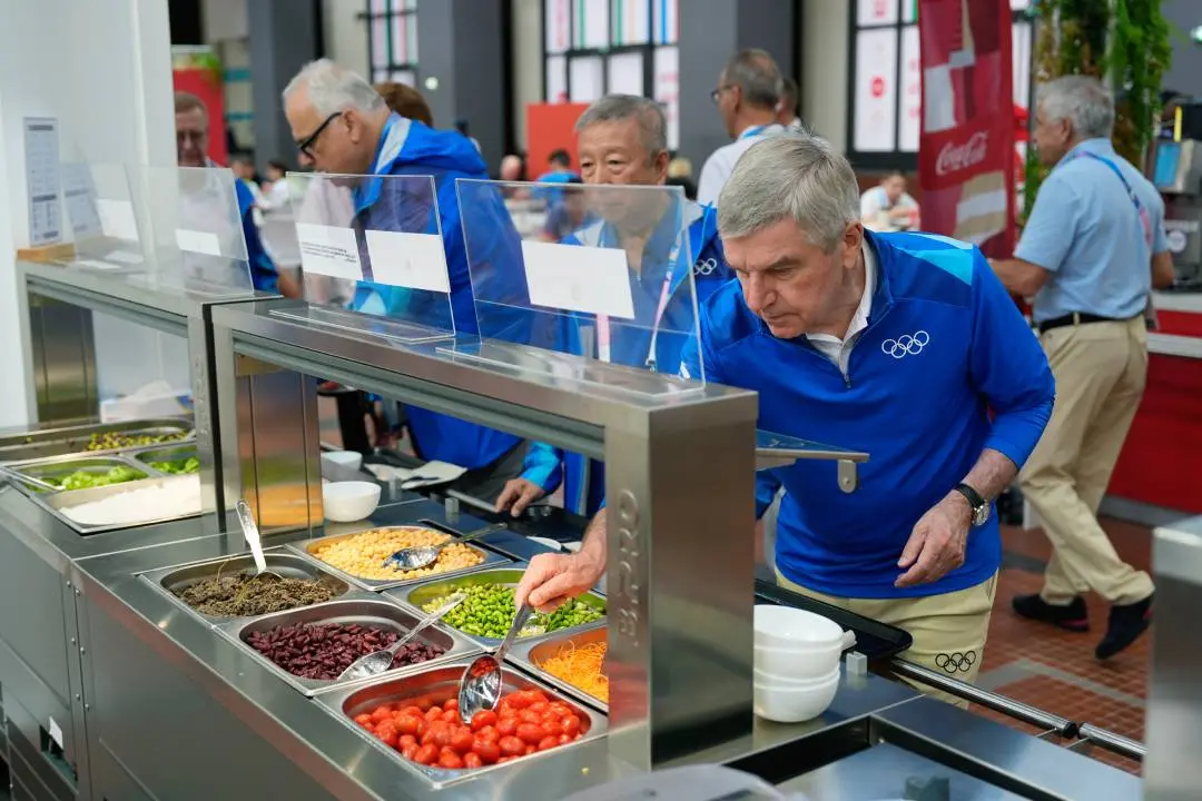 File photo (IOC President Thomas Bach tries food from a salad bar while touring the Olympic Village ahead of the 2024 Summer Olympics)
