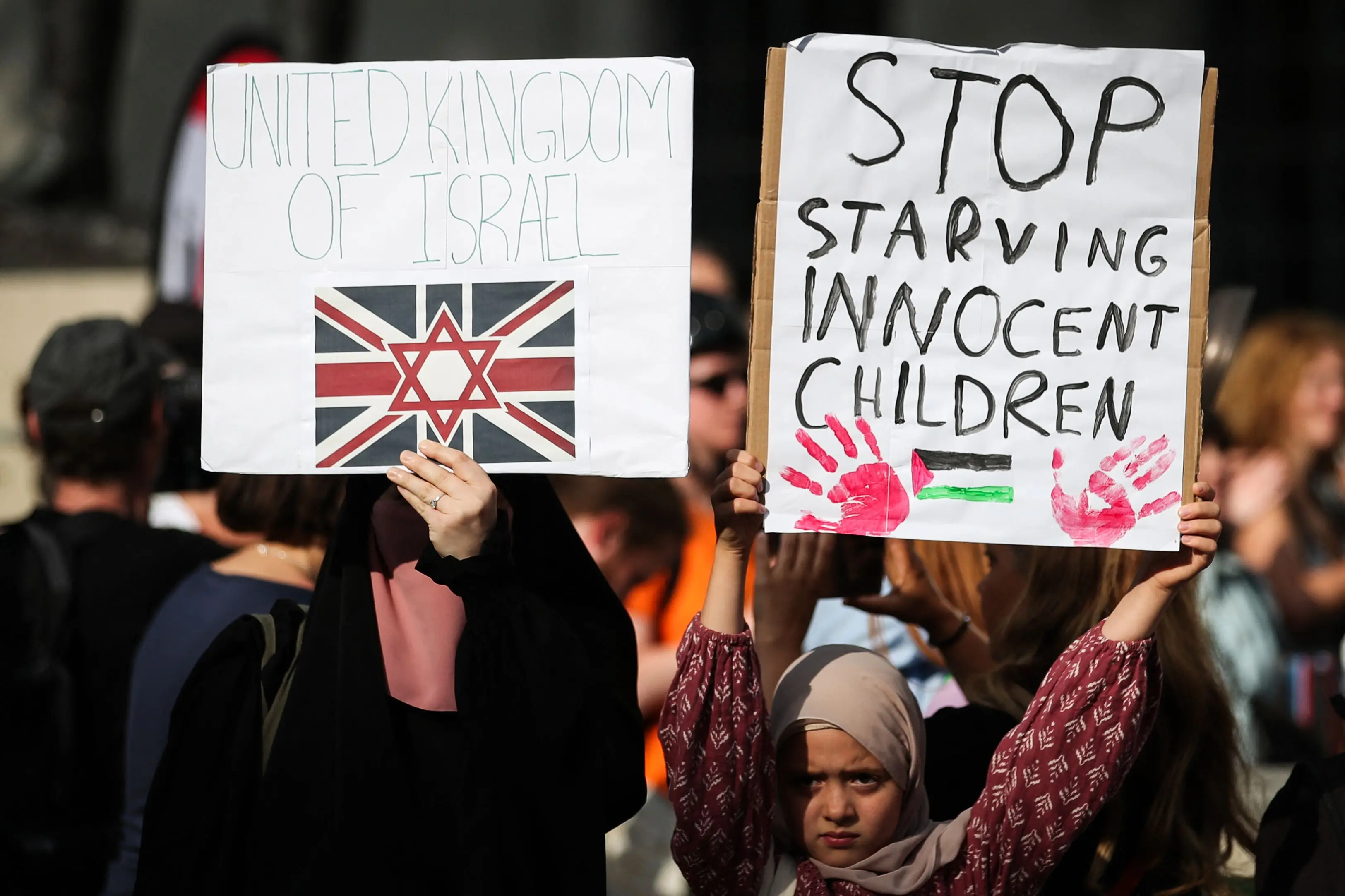 Protesters holds placards reading "United Kingdomof Israel" and "Stop starving innocent children" during a demonstration outside Downing Street gates, in central London. | Photo: AFP