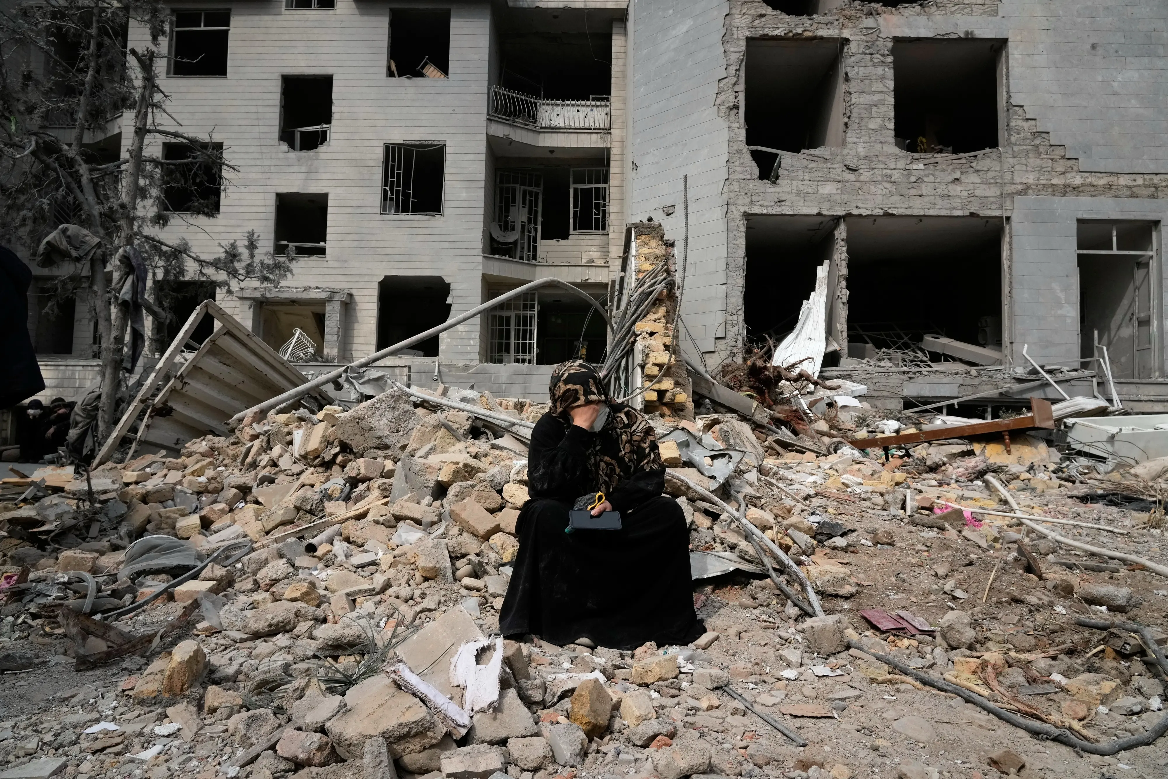 A woman sits on rubble across from a residential building damaged during the U.S.-Israeli air campaign in Tehran, Iran, March 12, 2026. (AP Photo/Vahid Salemi, File)
