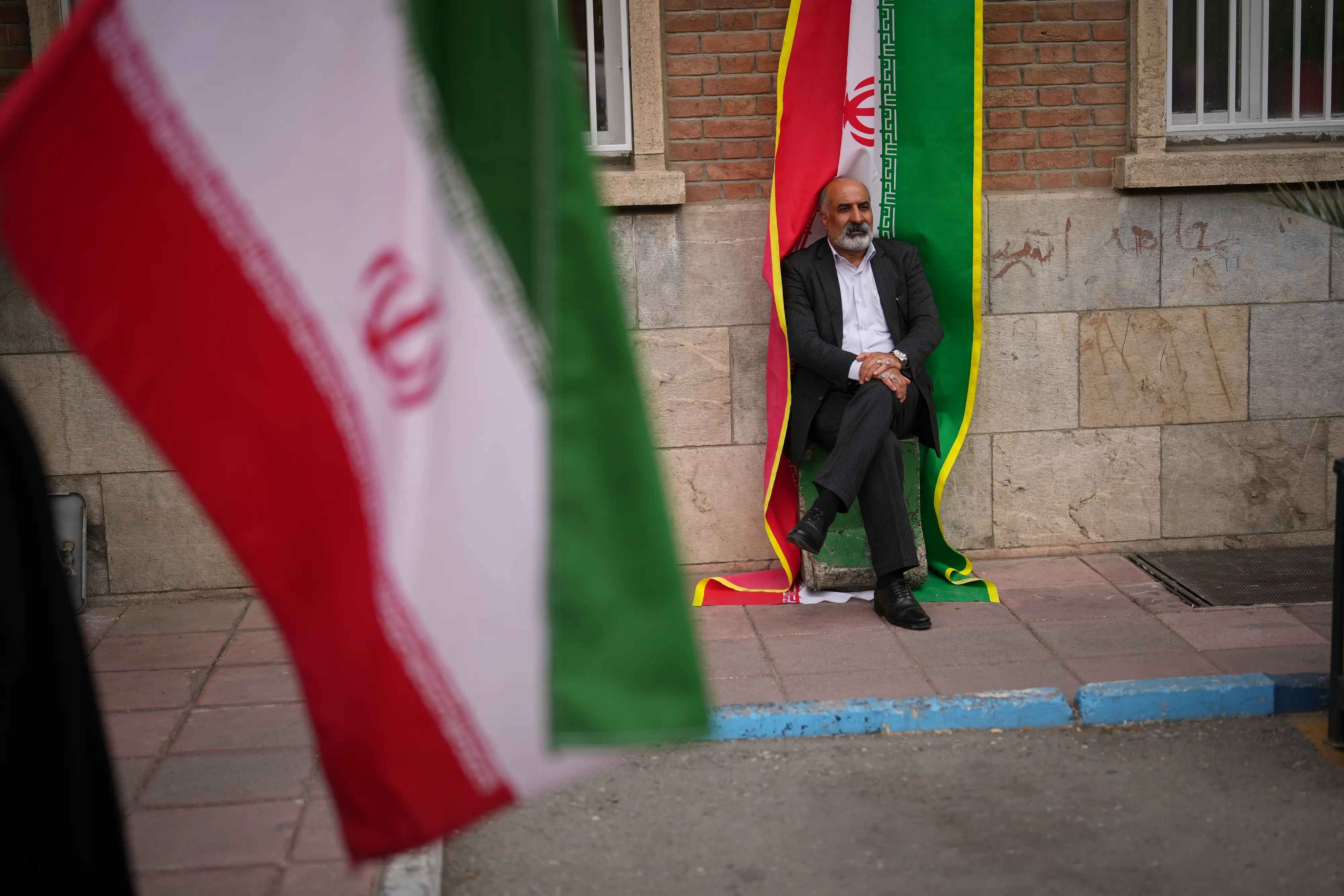 A man sits beside an Iranian flag banner during a government-sponsored protest attended by medical workers against the U.S.-Israeli military campaign outside Imam Khomeini Hospital in Tehran