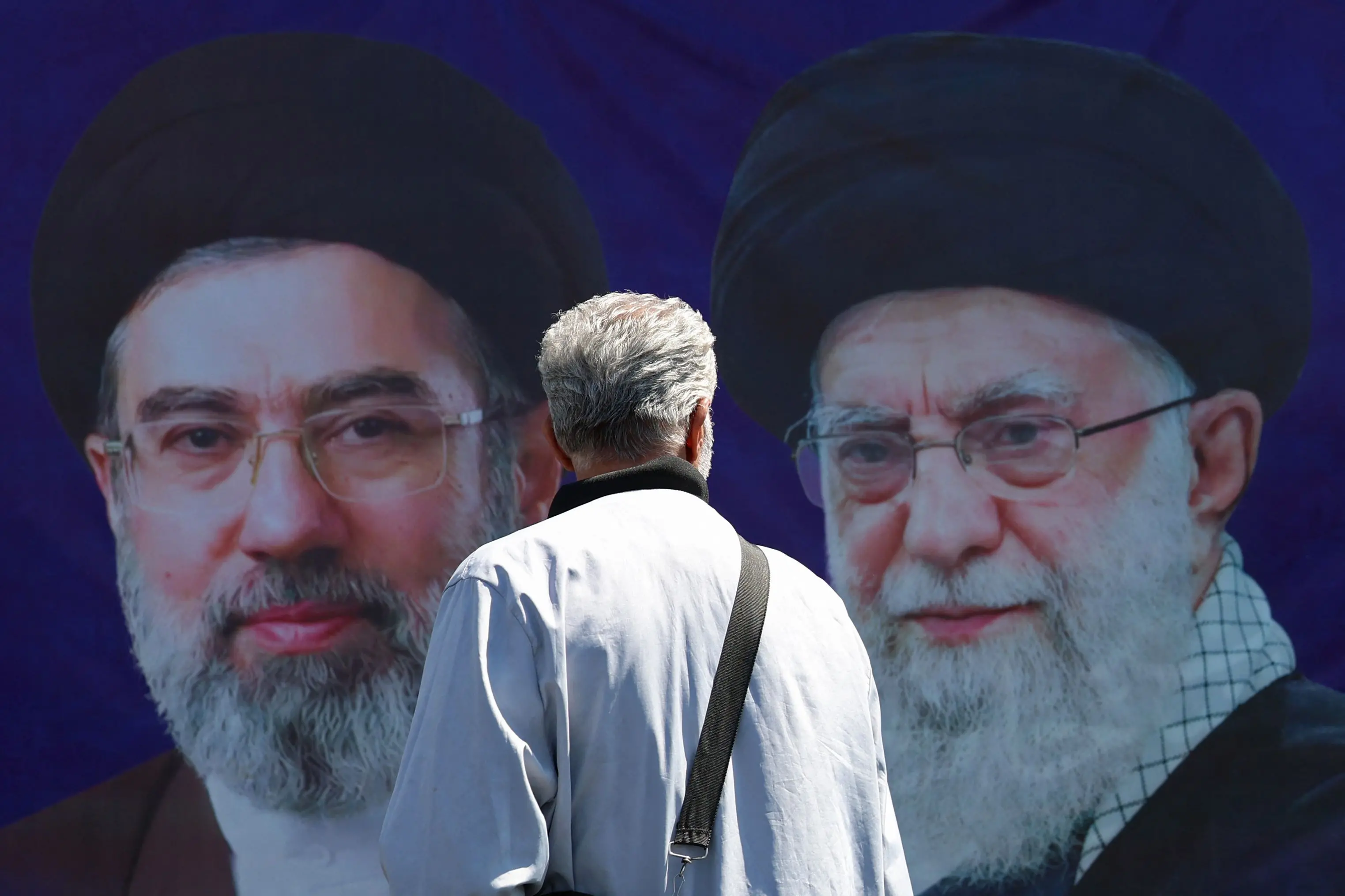 A man walks past a banner with pictures of Iran's slain supreme leader Ayatollah Ali Khamenei (R) and his son and successor Mojtaba Khamenei | AFP