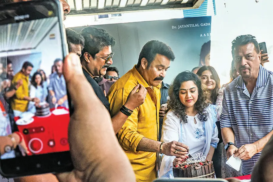 Malayalam actor Mohanlal cutting his birthday cake inside a crowded movie theatre surrounded by family, film crew, and cheering fans