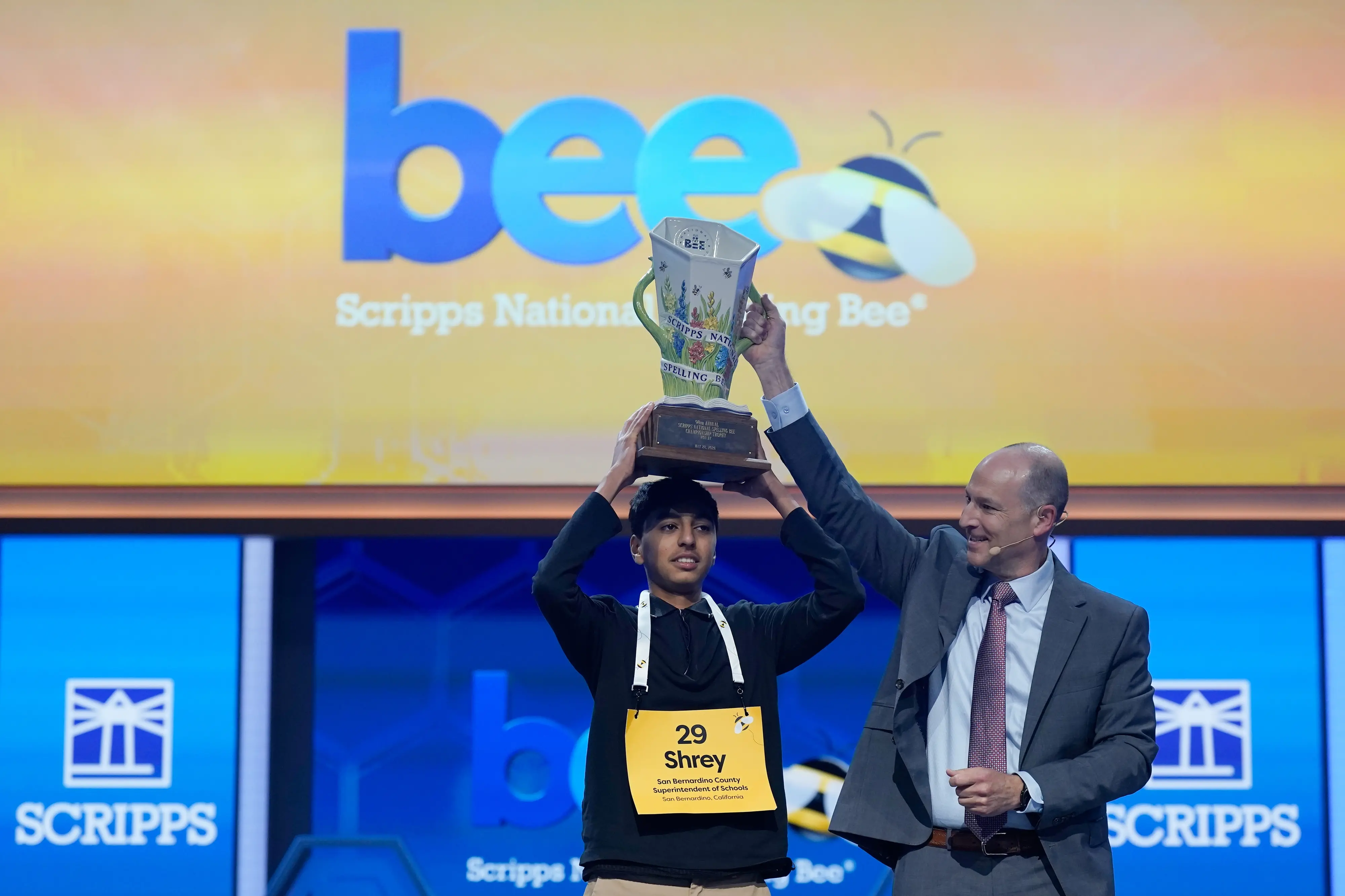 Scripps Company president and CEO Adam Symson, right, holds the trophy over winner of the 2026 Scripps National Spelling Bee, Shrey Parikh | AP